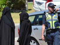 Two women stand next to a policeman blocking a road where police officers are searching a house in the Melbourne suburb of Meadow Heights