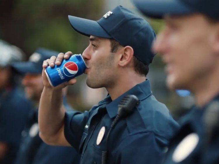 The crowd is heard cheering as a police officer drinks a can of Pepsi