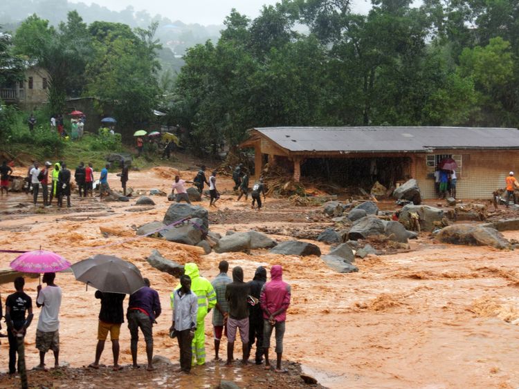 Bystanders look on as floodwaters rage past a damaged building in Freetown