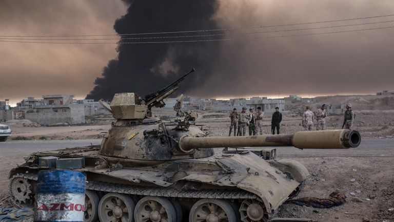 Iraqi soldiers stand next to a tank as smoke rises from the Qayyarah area