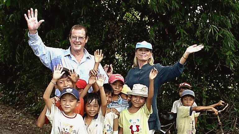 2003: Sir Roger Moore and his wife Kristina wave as they pose with children as part of a visit in his role as Goodwill Ambassador for the United Nations Children&#39;s Fund