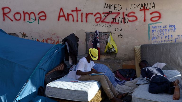 Migrants rest by a graffiti reading &#39;Rome antiracist&#39; in the street of Via Cupa outside the former Baobab migrant reception centre next to the Tiburtina train station in Rome on August 8, 2016. Set up almost three years ago the Baobab centre was shut down by police in December 2015 in the wake of Paris attacks. But Baobab volunteers quickly set up a camp on the street in front of the old shelter with tents and chemical toilets, serving three meals a day. / AFP / FILIPPO MONTEFORTE