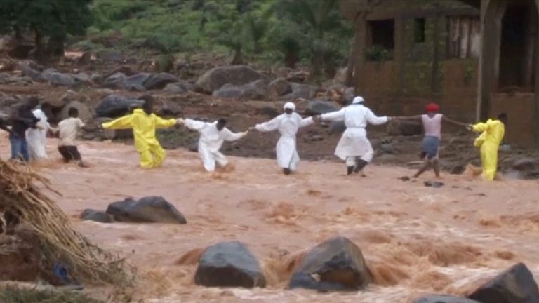 People wearing protective suits hold hands as they cross a river near one of the landslides