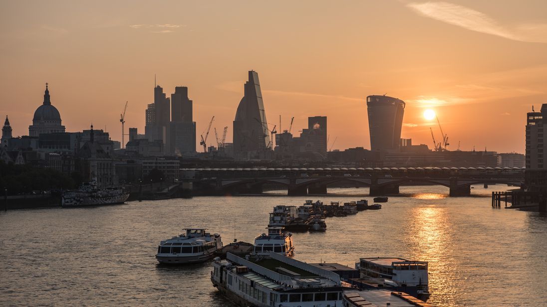 The sun rises over the City of London seen from Waterloo Bridge. Boats moored in the river Thames. The skyline includes St Paul's Cathedral, skyscrapers of The City including The Gherkin, the Cheesegrater, 20 Fenchurch Street 'Walki-Talkie'