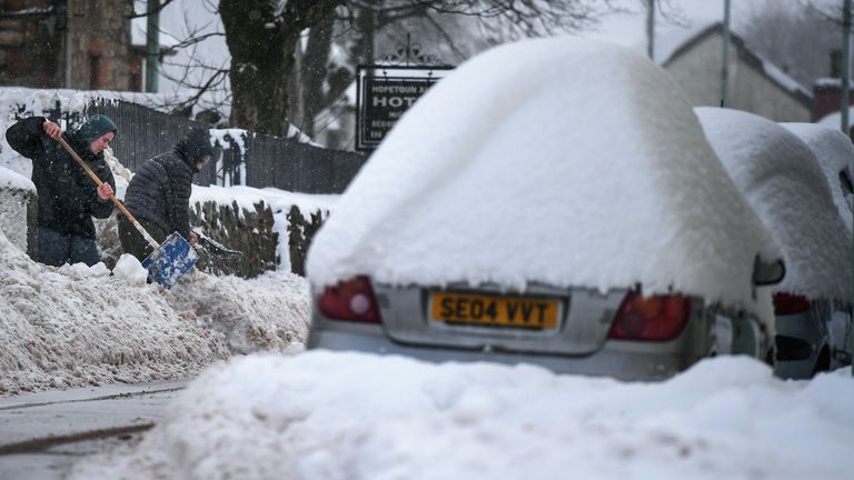 Locals try to clear snow from the front of their homes as parked cars sit covered in the white stuff in Dumfries, Scotland