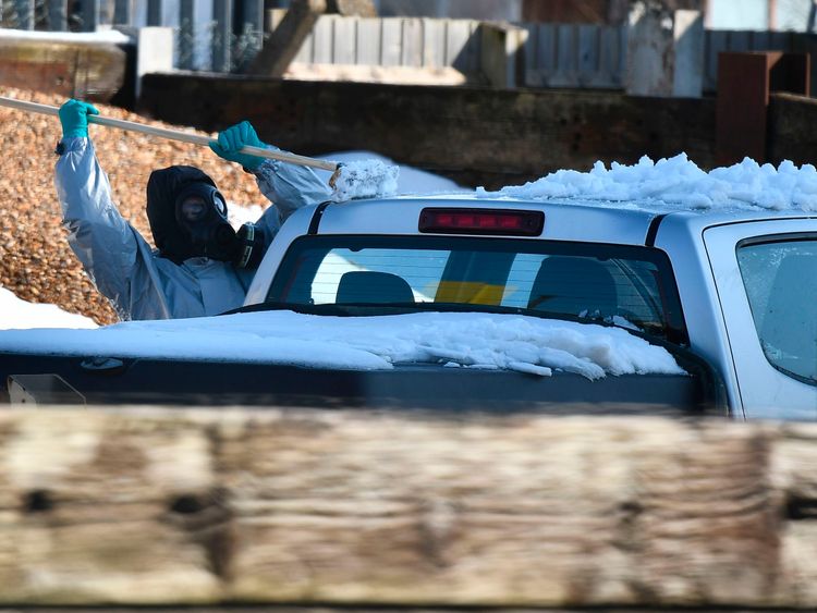 A worker in protective gear clears snow from a silver pick-up truck before removing it for examination