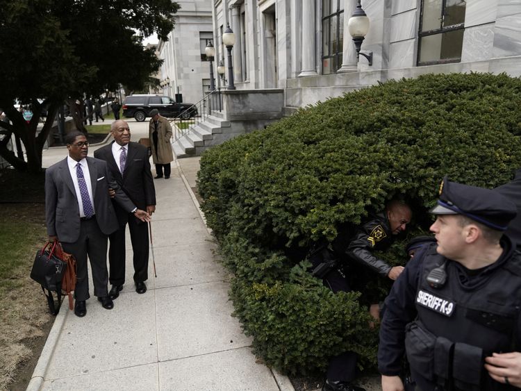 Bill Cosby watches as a protester struggles with police