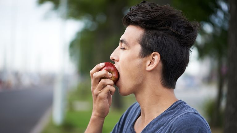 A young man taking a bite of an apple while waiting for the bus