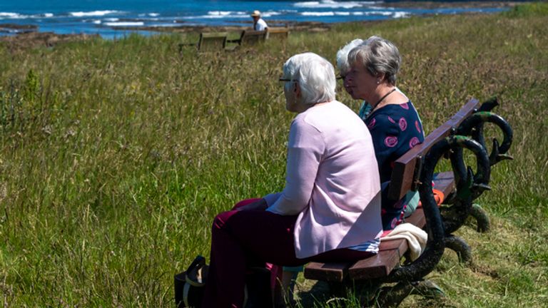 Three women looking out over the North Sea from the vantage point of a park bench looking towards the Farne islands with seahouses harbour in the background.