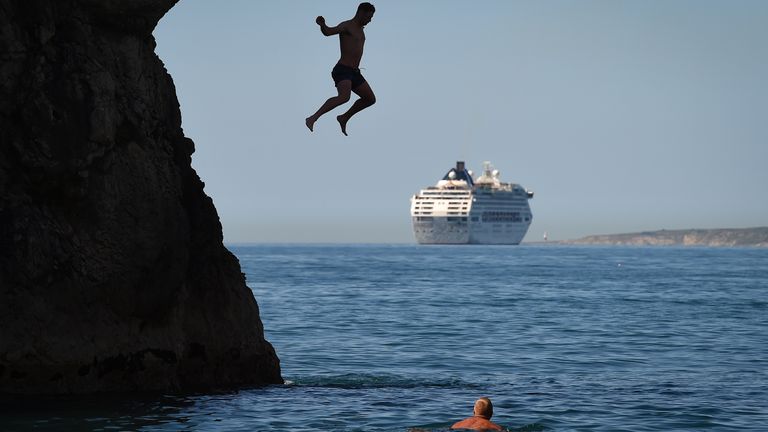 LULWORTH, ENGLAND - MAY 25: A man jumps from Durdle Door against the backdrop of a cruise ship as tourists enjoy the hot weather at Durdle Door beach on May 25, 2020 in West Lulworth, United Kingdom. The British government has started easing the lockdown it imposed two months ago to curb the spread of Covid-19, abandoning its 'stay at home' slogan in favour of a message to 'be alert', but UK countries have varied in their approaches to relaxing quarantine measures. (Photo by Finnbarr Webster/Get