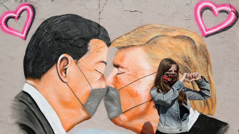 A young woman named Lily wears a face mask as she poses for her friend in front of graffiti of Communist Party of China General Secretary Xi Jinping (L) and U.S. President Donald Trump kissing each other while wearing face masks during the coronavirus crisis on April 27, 2020 in Berlin, Germany