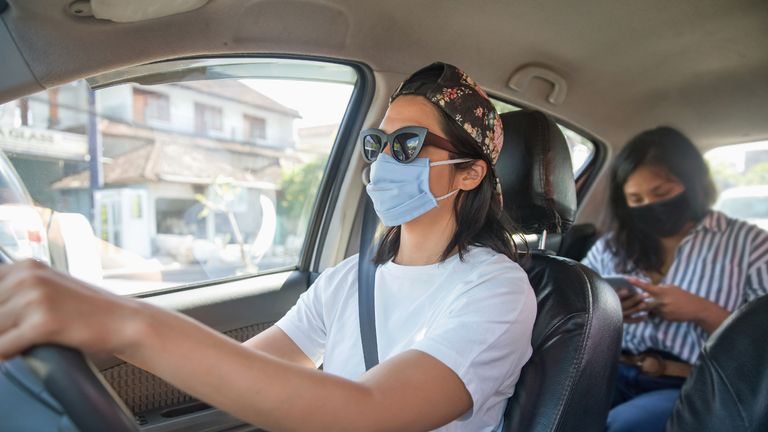 Portrait shot of mid adult Malaysian woman driving a car with a passenger behind. Both of the Malaysian woman are wearing protective face mask. The driver wearing a sunglasses and a cap. The passenger is a business woman who's busy with her smartphone.