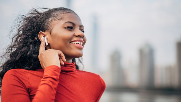 Woman standing downtown by the river and listening music on Airpod wireless headphones.