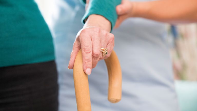 Senior Woman&#39;s Hands On Walking Stick With Care Worker In Background