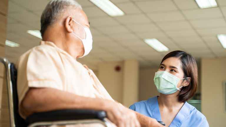 Asian nurse taking care of mature male patient sitting on wheelchair in hospital. Young woman and old man wearing surgical face mask for protection of The COVID pandemic. Girl talking to elderly man. (Asian nurse taking care of mature male patient sit