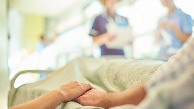 a hospital visitor&#39;s hand holds a patient&#39;s hand in bed of a hospital ward. In the blurred background a young nurse is chatting to the ward sister about the patient&#39;s care.