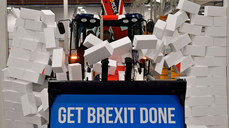 Britain's Prime Minister and Conservative party leader Boris Johnson drives a Union flag-themed JCB, with the words "Get Brexit Done" inside the digger bucket, through a fake wall emblazoned with the word "GRIDLOCK", during a general election campaign event at JCB construction company in Uttoxeter, Staffordshire, on December 10, 2019. - Britain will go to the polls on December 12, 2019 to vote in a pre-Christmas general election. (Photo by Ben STANSALL / POOL / AFP)