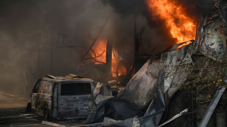 A burnt vehicle is seen as smoke and flames rise during a wildfire near the village of Afidnes, north of Athens