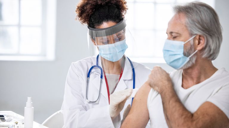Partial front view of mature Caucasian male patient watching as healthcare worker in lab coat, surgical mask, and face shield swabs injection site.