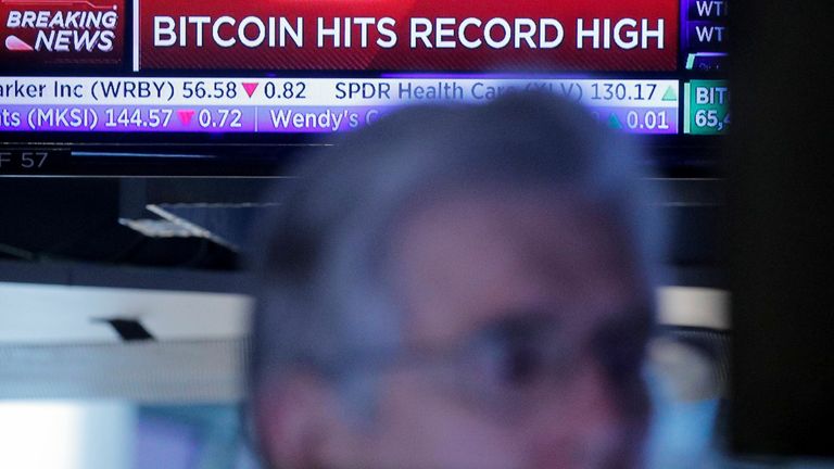 A screen displays a headline banner that reads; Bitcoin Hits Record High, as a trader works on the floor of the New York Stock Exchange