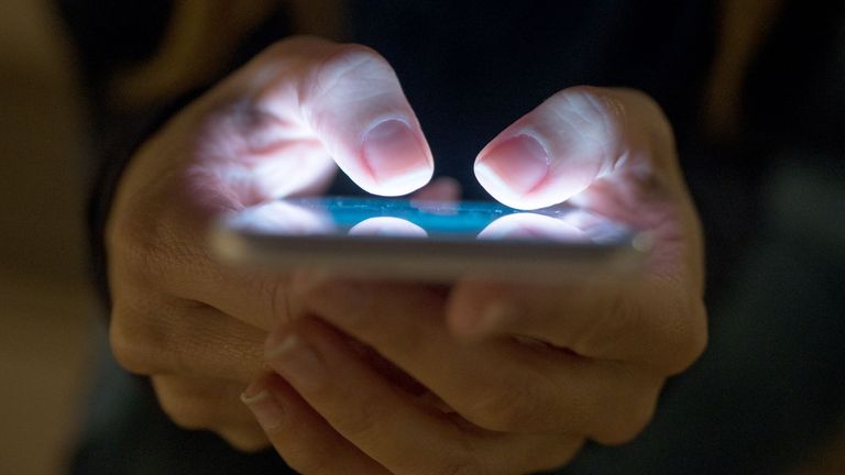 Front view of a woman pointing finger to smartphone with white monitor in the hands, working a computer keyboard on table. Pic: iStock