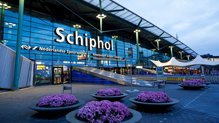 "Amsterdam, the Netherlands - October 14, 2012: The entrance of Schiphol Airport in Amsterdam in the evening with purple flowers in front of the airport hall. In the background some people standing outside."