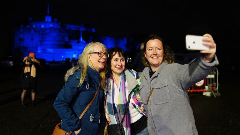 بازدیدکنندگان از لندن در مقابل قلعه ادینبورگ که به رنگ آبی برای هوگمانای روشن شده است سلفی می گیرند.