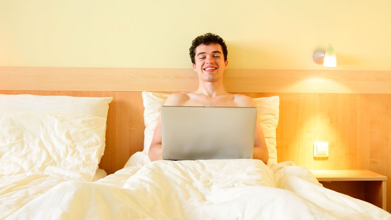 A pleased young man lying in bed with a laptop at his knees.


