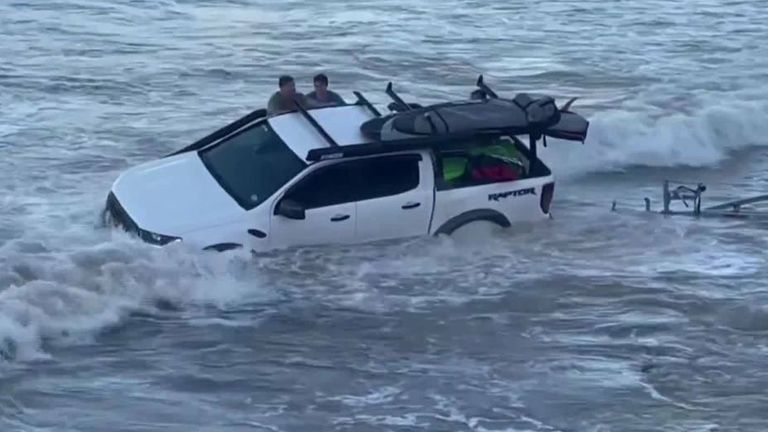 Strong waves caused by a cyclone rock a pick-up truck back and forth as surfers attempt to rescue it from the tide.