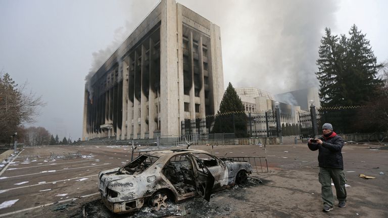 مردی با دیدن یک خودروی سوخته در مقابل ساختمان شهرداری که در اعتراضات ناشی از افزایش قیمت سوخت در آلماتی، قزاقستان، 6 ژانویه 2022 به آتش کشیده شد، عکس می گیرد. رویترز / پاول میخیف