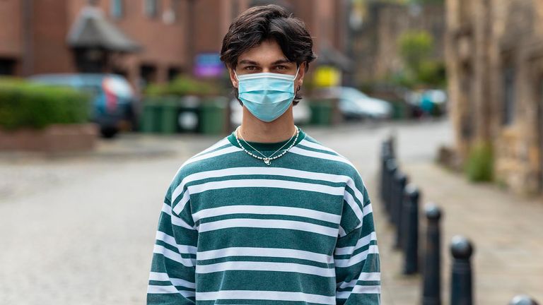 A portrait of a young man standing outside on a city street wearing a protective face mask while looking at the camera.  He protects himself from COVID-19.