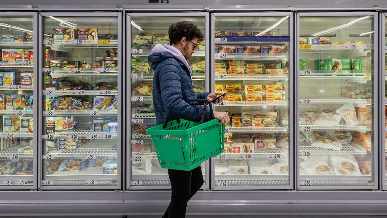 Man shopping in a supermarket while on a budget. He is looking for low prices due to inflation, standing looking at his phone in front of a row of freezers. He is living in the North East of England.

