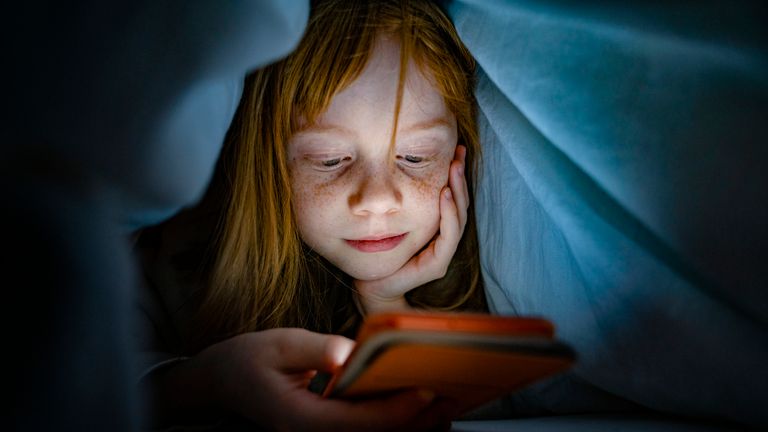 A girl uses a phone beneath a bed cover