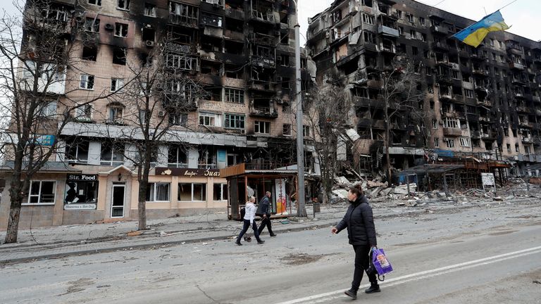 Ukrayna'nın güneyindeki liman kenti Mariupol'da Ukrayna-Rusya ihtilafı sırasında yıkılan bir apartmanın önündeki tele asılan yırtık Ukrayna bayrağının yakınında bir caddede yürüyen insanlar 14 Nisan 2022. REUTERS/Alexander Ermochenko