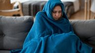 Young unsatisfied sick woman covered with warm blue blanket plaid from head to toes sitting alone on couch in living room at cold home. Seasonal problems, virus diseases and central heating concept