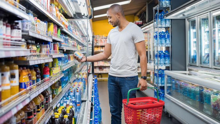 African man shopping in beverage section at supermarket. Black man doing shopping at market while buying cold drink. Handsome guy holding shopping basket reading nutritional values of product.