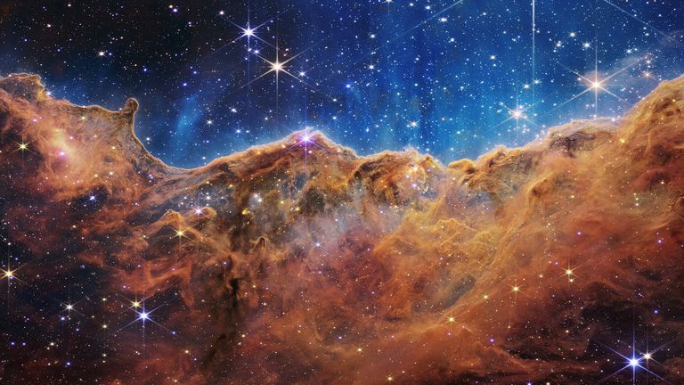 The "Cosmic Cliffs" of the Carina Nebula is seen in an image divided horizontally by an undulating line between a cloudscape forming a nebula along the bottom portion and a comparatively clear upper portion, with data from NASA&#39;s James Webb Space Telescope 