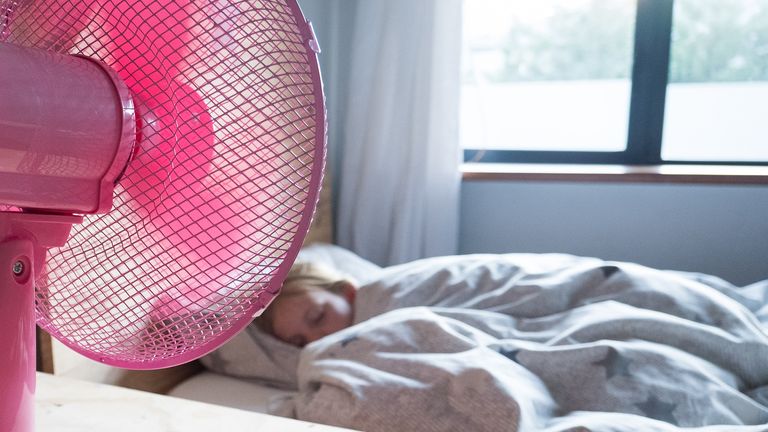 A young girl sleeps in her bed and has a pink fan on her desk blowing cool air over her.

