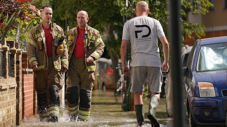 Anëtarët e Brigadës së Zjarrfikësve të Londrës kalojnë një pjesëtar të publikut teksa ecin në ujin e përmbytjes në Annette Road pranë Tollington Road, Holloway, në veri të Londrës, pas një shpërthimi të ujit prej 36 inç, duke shkaktuar përmbytje deri në katër këmbë të thellë. Data e fotografisë: e hënë 8 gusht 2022.