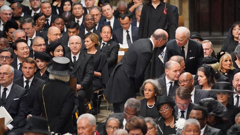 An attendant help Joe Biden find his seat, as dignitaries including Emmanuel Macron (left), looks on in contemplation