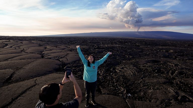 Kelly Ann Kobayashi raises her hands as she poses for a picture for Chad Saito, left, while standing on hardened lava rock from a previous eruption as the Mauna Loa volcano erupts, behind, Wednesday, Nov. 30, 2022, near Hilo, Hawaii. The two were visiting from Honolulu. (AP Photo/Gregory Bull)