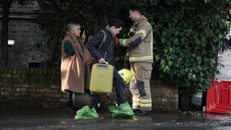 A firefighter talks to people with suitcases on Belsize Road in Camden after a burst water main flooded the London street. London Fire Brigade (LFB) said eight fire engines and around 60 firefighters were called to Belsize Road at 2.50am on Saturday morning after a 42-inch water main burst, causing flooding to a depth of half a metre across an area of around 800 metres. Picture date: Saturday December 17, 2022.

