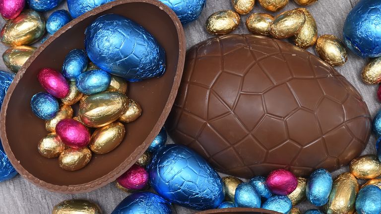 Pile or group of multi colored and different sizes of colourful foil wrapped chocolate easter eggs in pink, blue, and gold. Large halves of a brown milk chocolate egg have mini eggs inside, on a grey wooden background. 