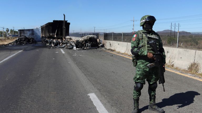 A soldier stands guard near the wreckage of a burnt out vehicle set alight by drug gang members as a barricade