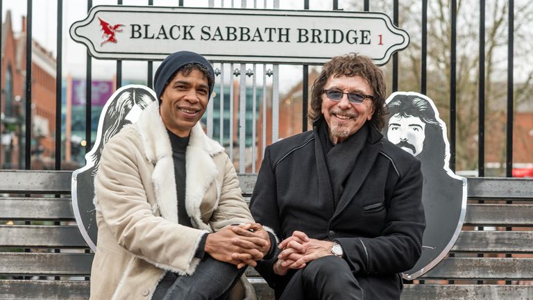 Director of Birmingham Royal Ballet, Carlos Acosta, and Black Sabbath guitarist, Tony Iommi, sat in front of a sign for Black Sabbath Bridge in Birmingham.