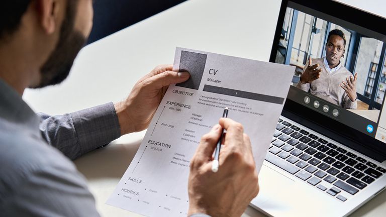 Recruiter or employer with resume in hand conducts online virtual job interview with candidate via video call. Remote remote recruitment meeting chat. Over the shoulder view. Figure: iStock