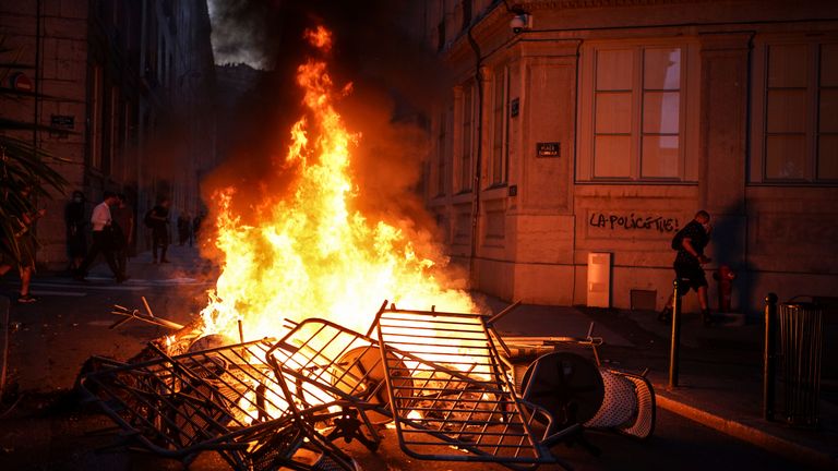 Bir adam, üzerinde 'Polis öldürür' yazan yanan bir barikatın aydınlattığı bir duvarın yanından geçer.  Fransa'nın merkezinde, Lyon'un merkezinde, 30 Haziran 2023 Cuma. Fransa Cumhurbaşkanı Emmanuel Macron, Cuma günü ebeveynleri gençleri evde tutmaya çağırdı ve 17 yaşındaki bir gencin polis tarafından öldürülmesi üzerine Fransa genelinde yayılan isyanı bastırmak için sosyal medyada kısıtlamalar önerdi. -eski sürücü.  Duvarda Fransızca 