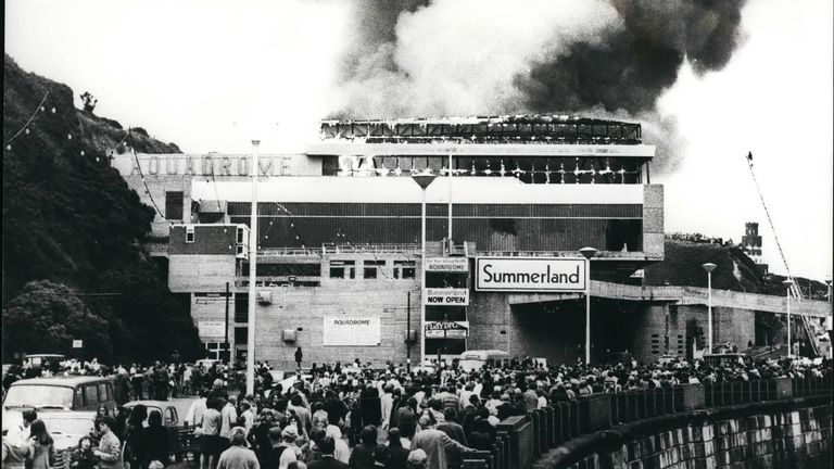 Aug. 08, 1973 - 42 die in the Isle of Man Fun Center Holiday Blaze: The death toll in the Isle of Man holiday blaze rose to 41 today and there are fears that the final figure would reach 50. Firemen continued their search of the ruins which had once been the £2 million Summerland fun center at Douglas, the island&#39;s capital. The blaze last night was at the peak of the Island&#39;s holiday season. Photo shows Holiday makers look on as clouds of smoke and flames pour out from the Summerland fun c