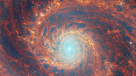  A large spiral galaxy takes up the entirety of the image. The core is mostly bright white, but there are also swirling, detailed structures that resemble water circling a drain. There is white and pale blue light that emanates from stars and dust at the core’s centre, but it is tightly limited to the core. The rings feature colours of deep red and orange and highlight filaments of dust around cavernous black bubbles.