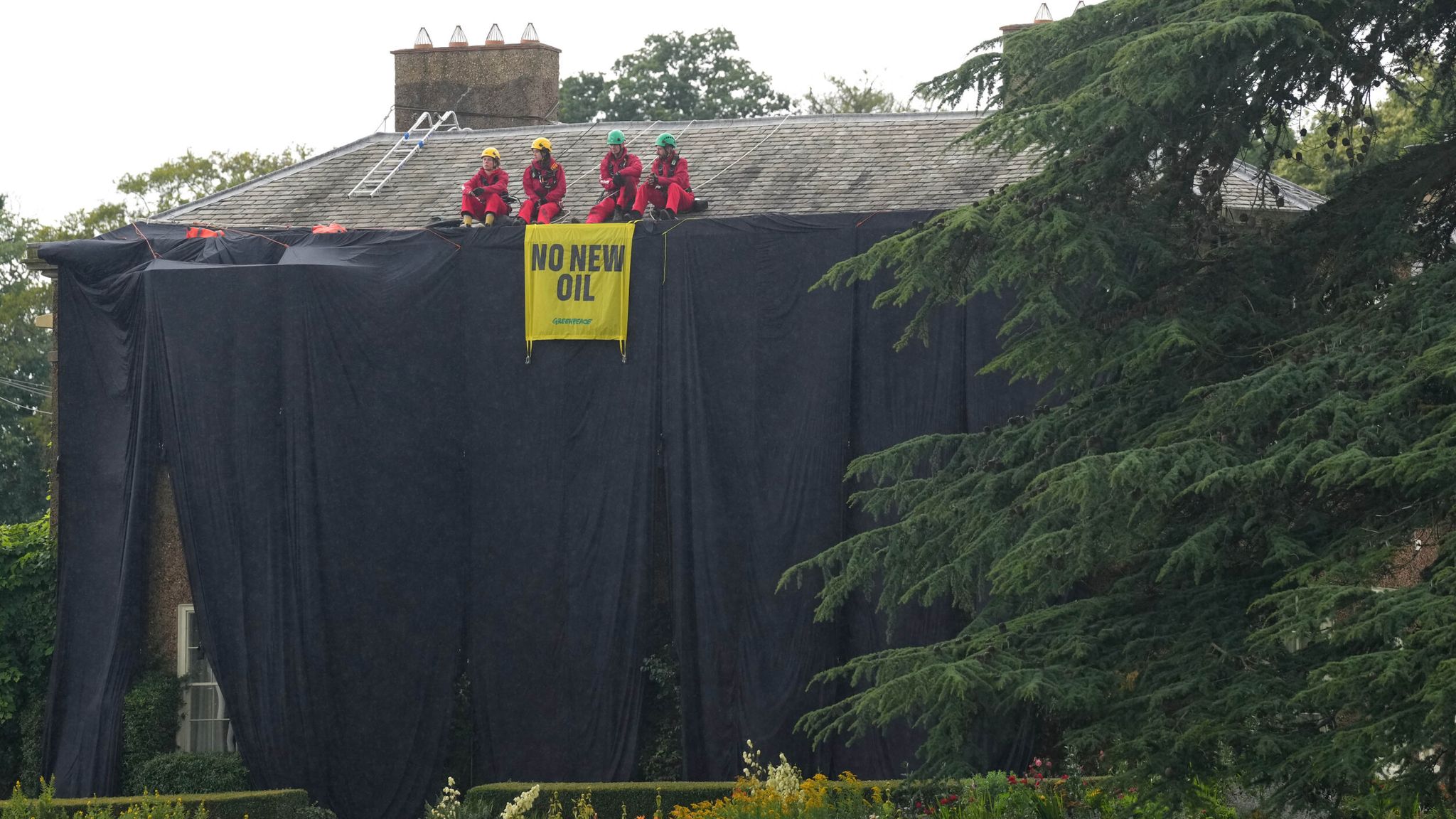 Four protesters in red safety gear sit on a house roof, displaying a yellow 